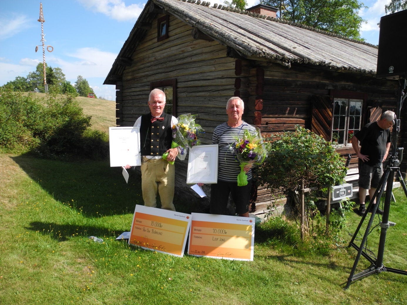 Två män står i gräset utanför knuten på en fjällstuga. Båda håller i varsin blombukett och en tavelram med diplom.