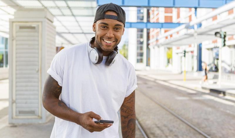 Smiling young man with headphones and smartphone waiting at tram stop